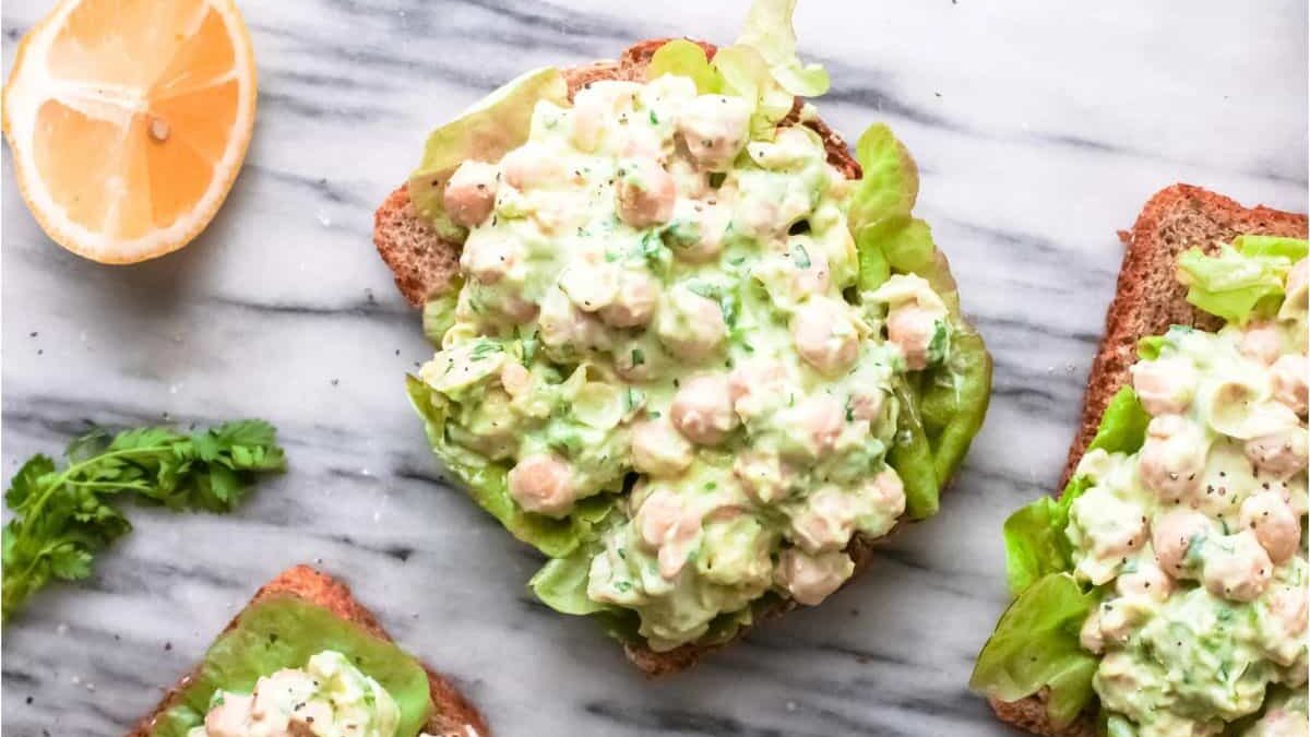 Open-faced sandwiches on a marble surface, topped with a creamy chickpea salad and lettuce. A lemon wedge and fresh parsley are nearby.