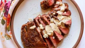 A sliced, medium-rare steak topped with creamy peppercorn sauce and blue cheese crumbles on a white plate with a brown rim, next to a floral patterned napkin.