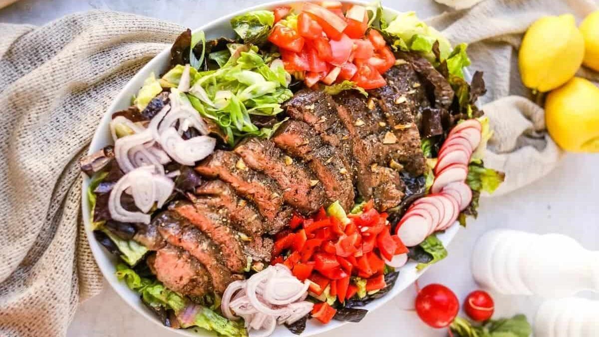 A platter of sliced grilled steak served over a bed of mixed greens, topped with chopped tomatoes, radishes, onions, and red bell peppers, with lemons and vegetables on the side.