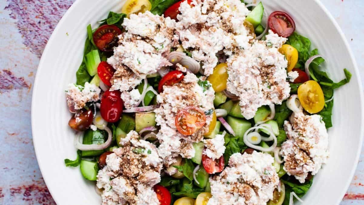A colorful salad on a white plate, featuring mixed greens, cherry tomatoes, cucumber slices, and dollops of ricotta. It's garnished with a sprinkle of black pepper and thinly sliced rings of red onion.