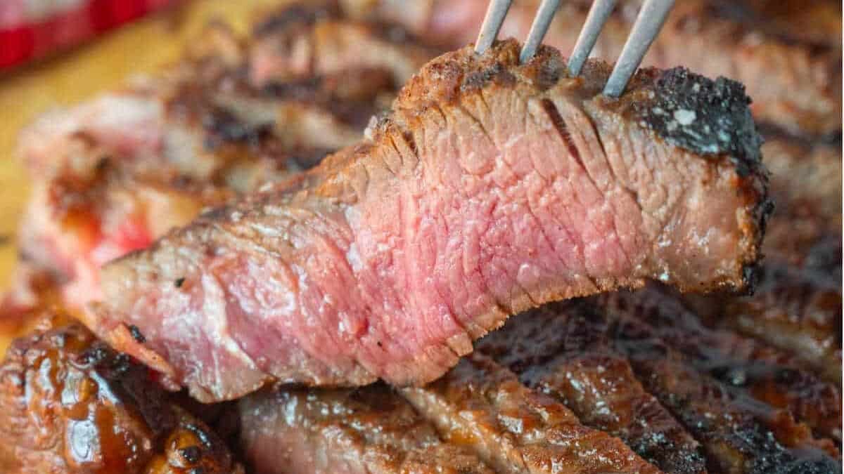 A close-up of a juicy, sliced steak cooked medium-rare, with a fork holding up one slice. The meat is browned on the outside and pink in the center, showing a tender, grilled texture.