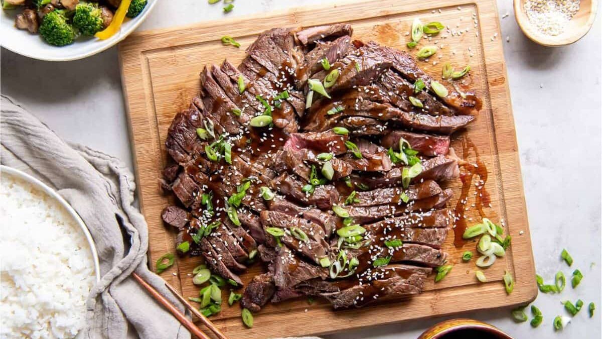 Sliced grilled steak topped with sesame seeds and chopped green onions on a wooden board, surrounded by bowls of rice, vegetables, and sauce, with chopsticks on the side.