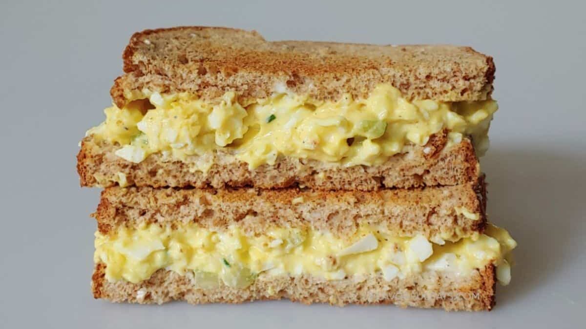A sandwich made with whole grain bread is cut in half and stacked. It is filled with creamy egg salad, which includes chopped eggs and herbs. The filling is spilling slightly from the sides. The background is a plain, neutral surface.