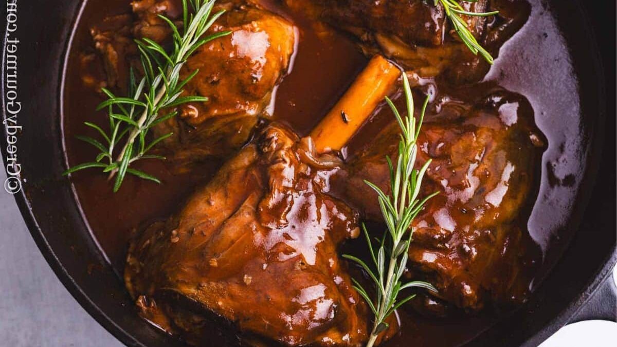 A close-up of a cast iron skillet filled with braised lamb shanks in a rich brown sauce, garnished with fresh rosemary sprigs.