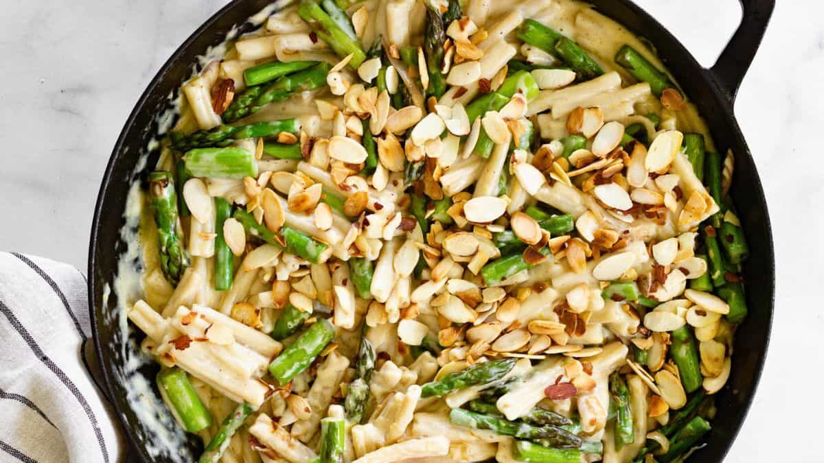 A skillet filled with creamy pasta features asparagus and sliced almonds on top. A wooden spoon rests inside the dish, atop a white and black striped cloth, on a marble surface.