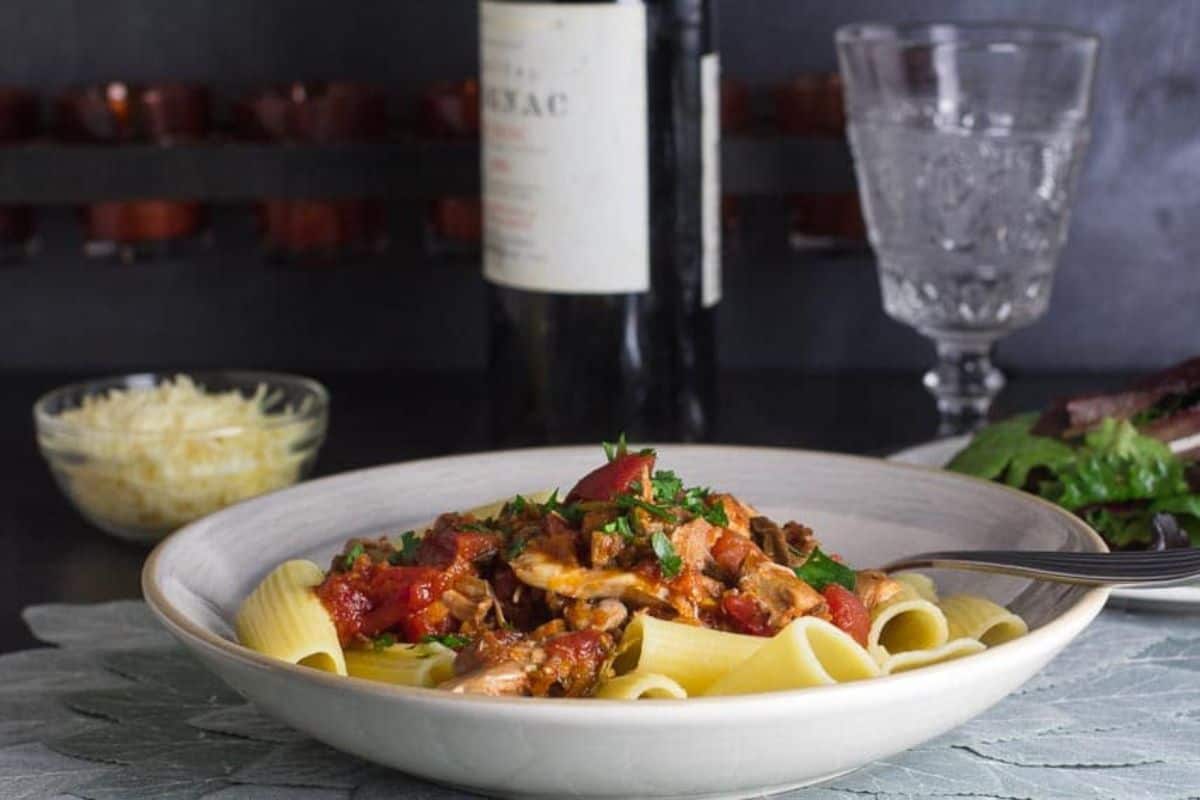 A bowl of pasta topped with a rich tomato and mushroom sauce, garnished with herbs. Accompanied by a side salad and a glass of wine, with a bottle in the background. A bowl of grated cheese sits nearby on a textured tablecloth.