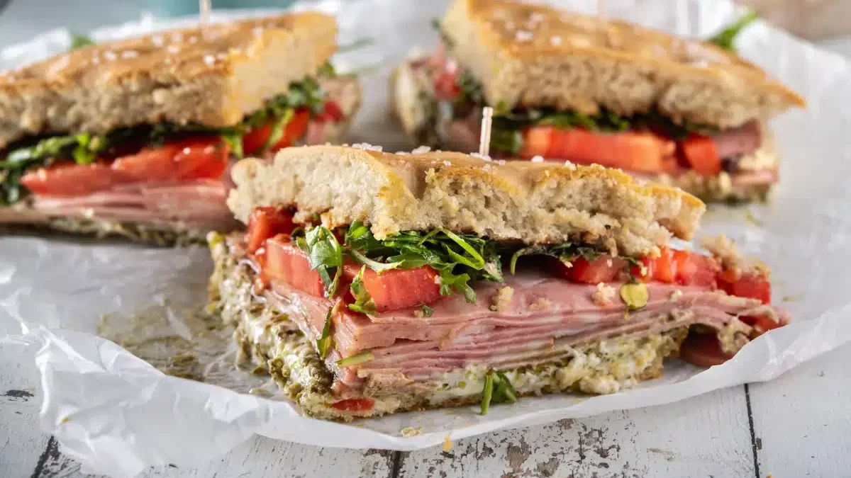 Close-up of a sliced sandwich with layers of ham, fresh tomatoes, greens, and spread between rustic bread. The sandwich is on crumpled parchment paper on a wooden surface.