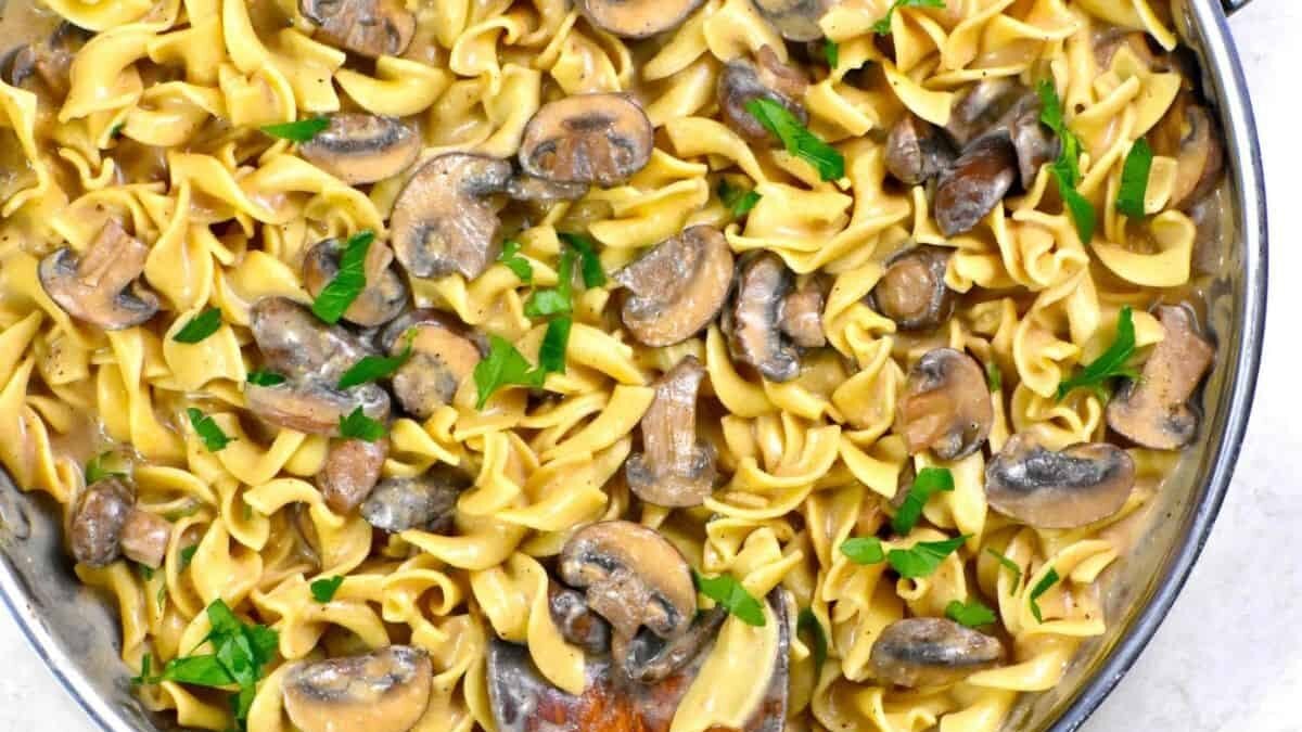 A pan filled with mushroom stroganoff, featuring wide egg noodles, sliced mushrooms, and a creamy sauce. Garnished with fresh parsley, and a wooden spoon is partially visible in the dish.