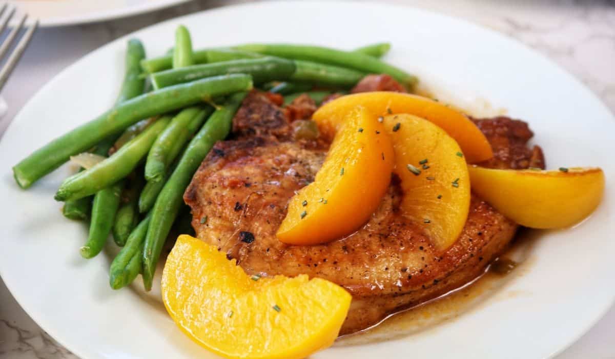 A white plate with a seasoned pork chop topped with orange slices, served alongside cooked green beans.