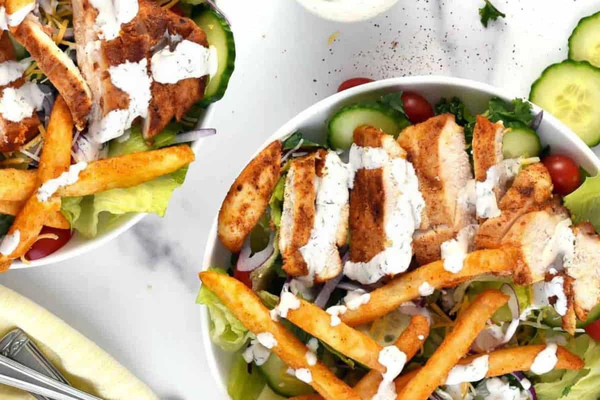 Two bowls of salad with grilled chicken slices, cherry tomatoes, fries, and cucumber slices, topped with creamy dressing. The salads are served on a white surface with additional cucumber slices nearby.