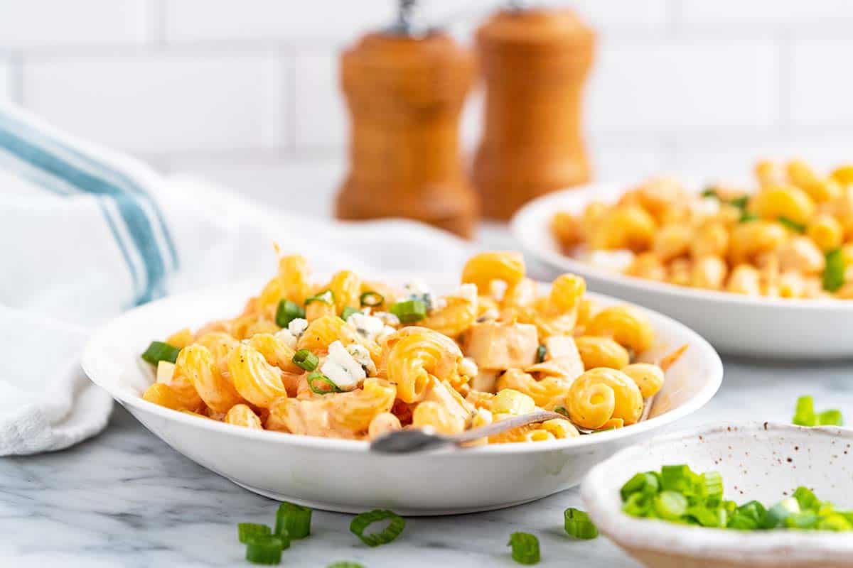 A bowl of creamy pasta with chopped green onions and cheese crumbles, set on a marble countertop. A second bowl and wooden salt and pepper shakers are blurred in the background. A striped kitchen towel is to the side.