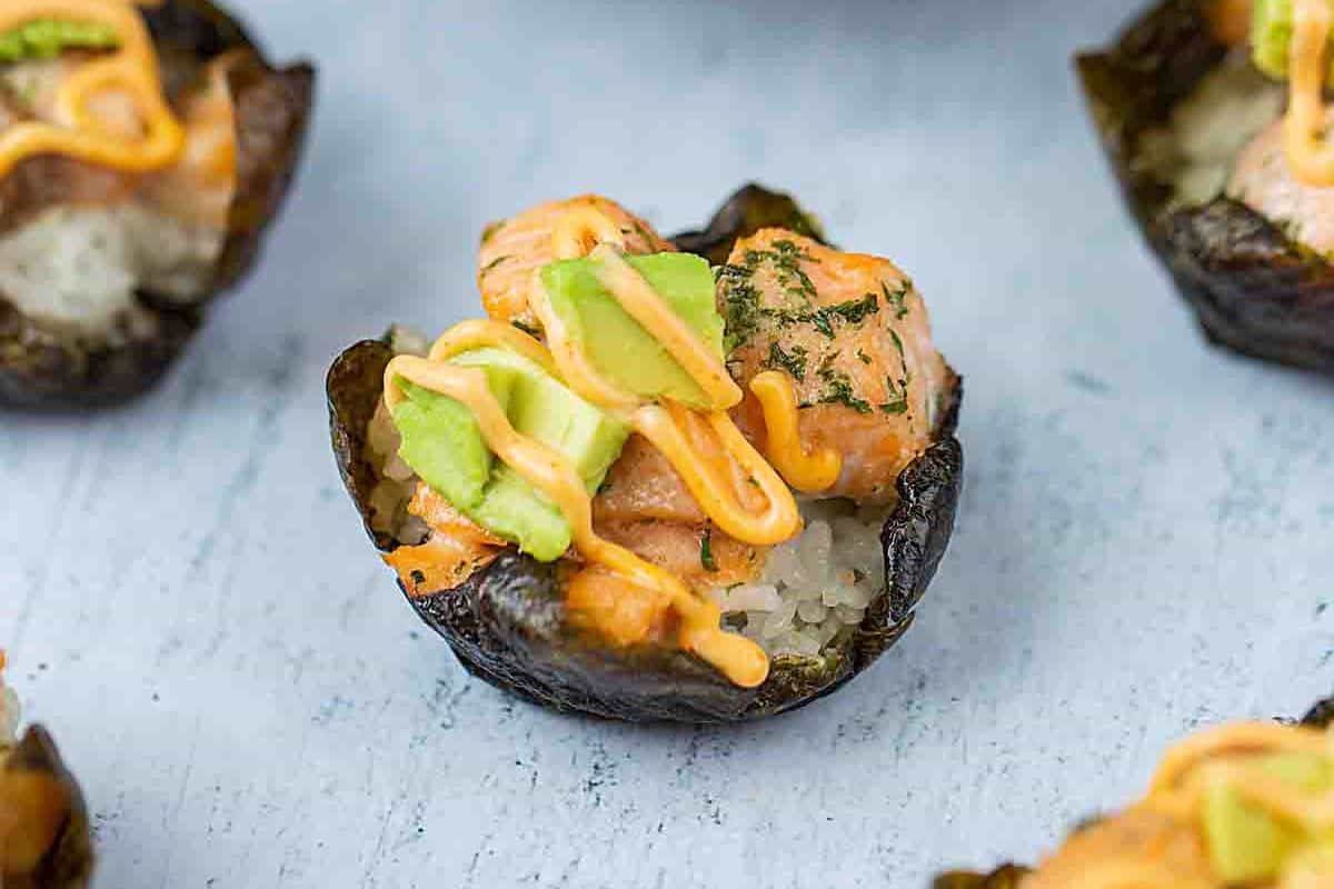 A bite-sized sushi cup with a nori (seaweed) wrap, filled with rice, chunks of salmon, avocado slices, and drizzled with a spicy orange sauce. The sushi cup is placed on a light gray surface.