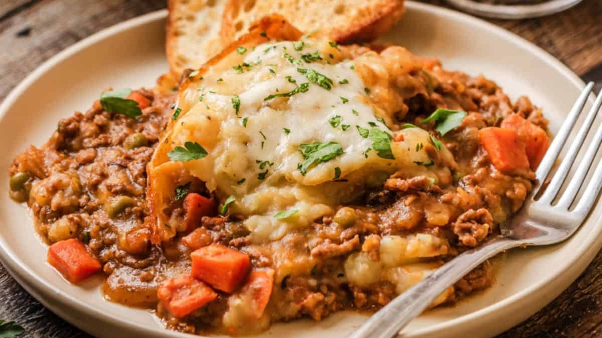 A plate of shepherd's pie with a golden mashed potato topping, served alongside carrot and peas in a rich gravy. Two slices of toasted bread are in the background, with a fork resting on the plate.