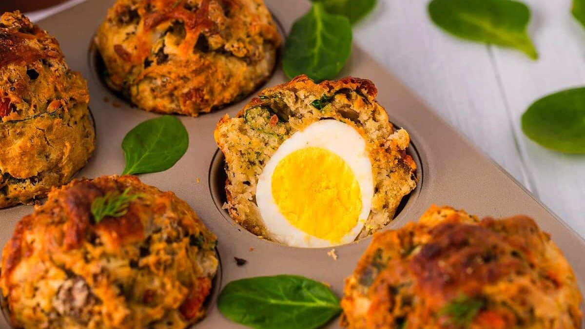 Muffin tin containing baked muffins with spinach and whole boiled eggs inside. A muffin is cut in half to reveal the egg. Spinach leaves are scattered on a wooden surface.