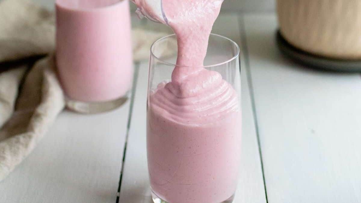A thick pink smoothie is being poured into a clear glass, with another glass of pink smoothie in the background on a white surface.