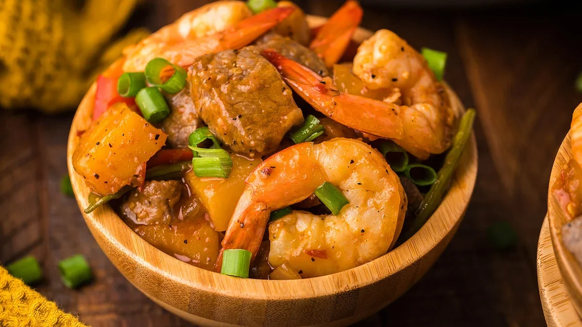 A wooden bowl filled with a colorful dish featuring shrimp, beef chunks, pineapple pieces, sliced red bell peppers, green onions, and a savory sauce. The food is garnished with chopped green onions.