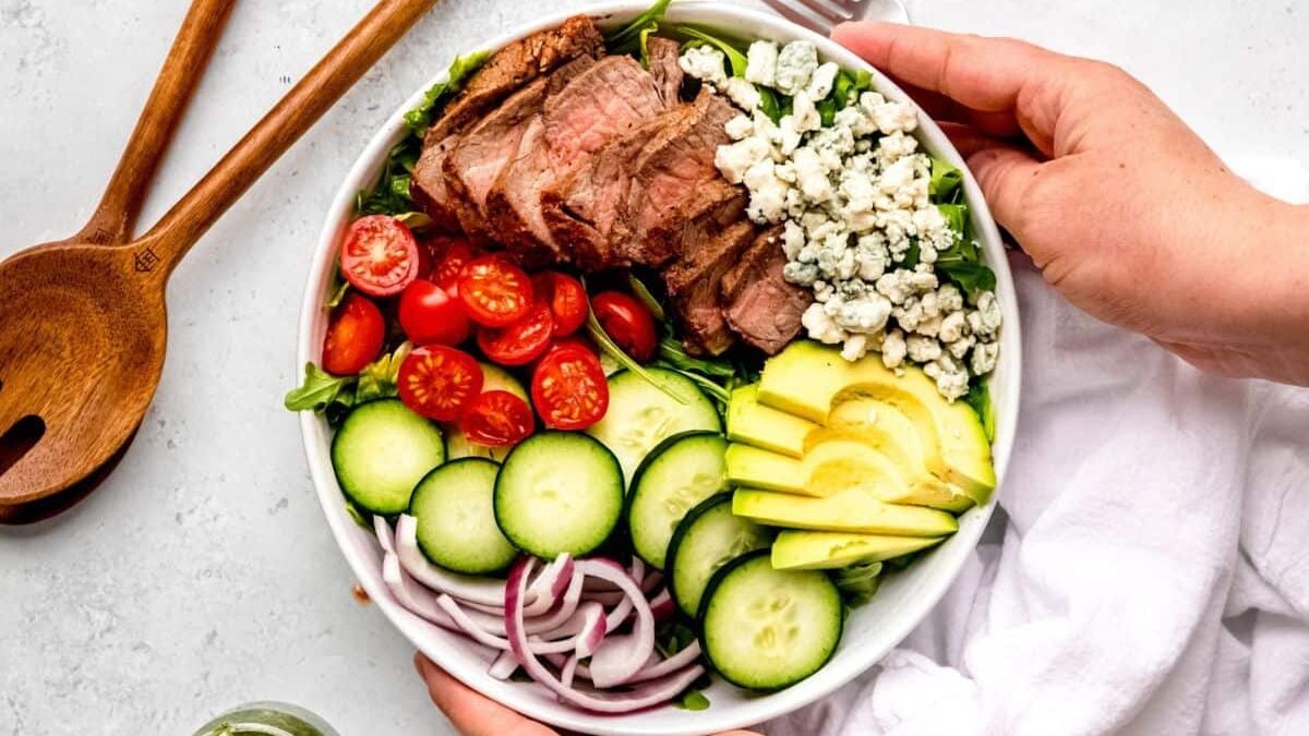 A bowl of salad with sliced steak, cherry tomatoes, cucumber, red onion, avocado, crumbled blue cheese, and greens. Two hands hold the bowl, and wooden salad utensils are nearby on a white surface.
