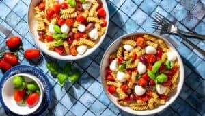 Two bowls of rotini pasta salad with cherry tomatoes, mozzarella balls, black olives, and fresh basil leaves. A small bowl with cherry tomatoes and basil is next to them on a blue tiled surface. Two forks lie beside the bowls.