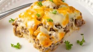 A slice of cheesy beef casserole on a white plate, topped with melted cheddar and mozzarella cheese, garnished with fresh parsley. A fork rests beside the slice.