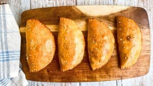 Four golden-brown empanadas with sesame seeds are lined up on a wooden cutting board, next to a blue and white checkered cloth.