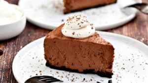 A slice of chocolate cheesecake on a white plate, topped with a swirl of whipped cream and sprinkled with chocolate shavings. A fork is nearby, and another dessert plate with a similar slice is blurred in the background.