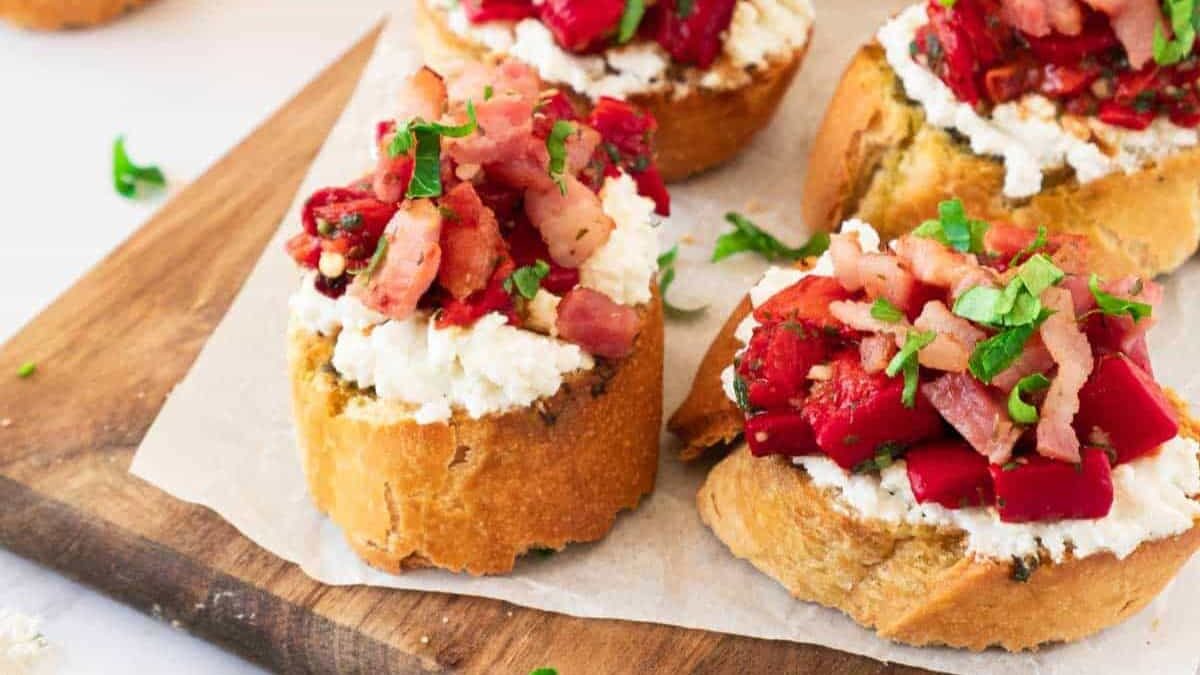 Slices of bread topped with white cheese, diced tomatoes, and herbs are arranged on a wooden board lined with parchment paper.