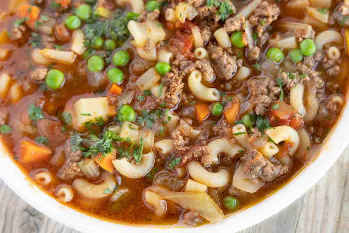 A close-up of a bowl of hearty soup with ground beef, elbow macaroni, green peas, diced carrots, and chopped herbs in a rich, tomato-based broth.