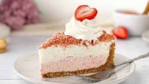 A slice of strawberry cheesecake with a graham cracker crust, topped with whipped cream and a fresh strawberry slice, is placed on a white plate. The background is softly blurred with other strawberries and pink flowers visible.