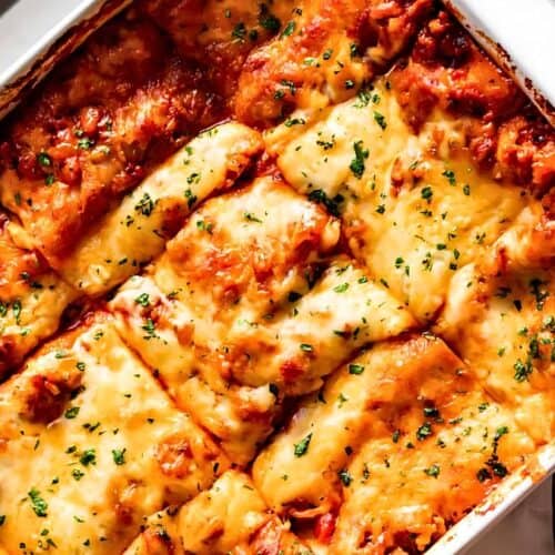 A close-up of a baked lasagna in a rectangular dish, with golden melted cheese, tomato sauce, and sprinkled herbs on top. A knife and striped towel are next to the dish.