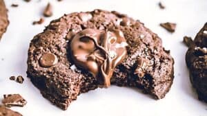 A close-up of a chocolate cookie with a gooey chocolate center and chocolate chunks, shown with a bite taken out, on a white background.
