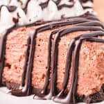 A close-up of a slice of chocolate mousse pie on a white plate. The pie has a chocolate crust, creamy mousse filling, and is topped with whipped cream and drizzled with chocolate syrup. A fork is placed next to the slice.