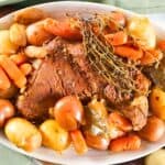 A platter featuring a cooked pot roast, surrounded by carrots and various types of potatoes, garnished with herbs. It rests on a pale green napkin with additional vegetables and bread nearby.