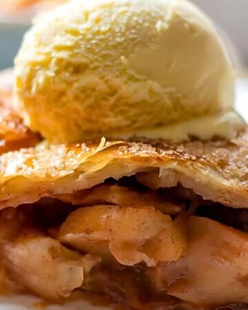 A slice of apple pie on a plate, topped with a scoop of vanilla ice cream. The pie has a golden, flaky crust and visible cinnamon-spiced apple filling.