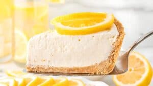 A slice of creamy lemon pie with a crumbly crust, garnished with lemon slices, is being lifted from a pie dish. In the background, there are more lemon slices and a blurred setting with yellow tones.