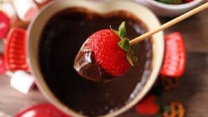 A skewer holding a strawberry covered in chocolate over the fondue pot.