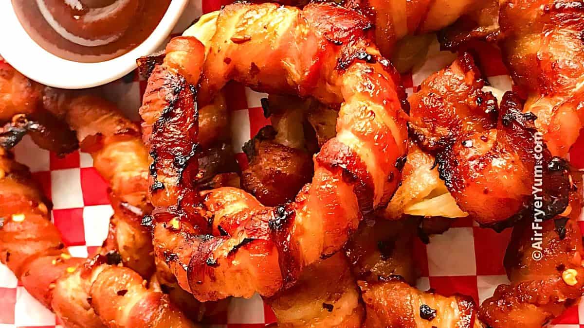Close-up of crispy, bacon-wrapped onion rings on a red and white checkered serving tray. A small dish of dipping sauce is partially visible in the top left corner. The onion rings are charred and well-seasoned, displaying a golden-brown color.