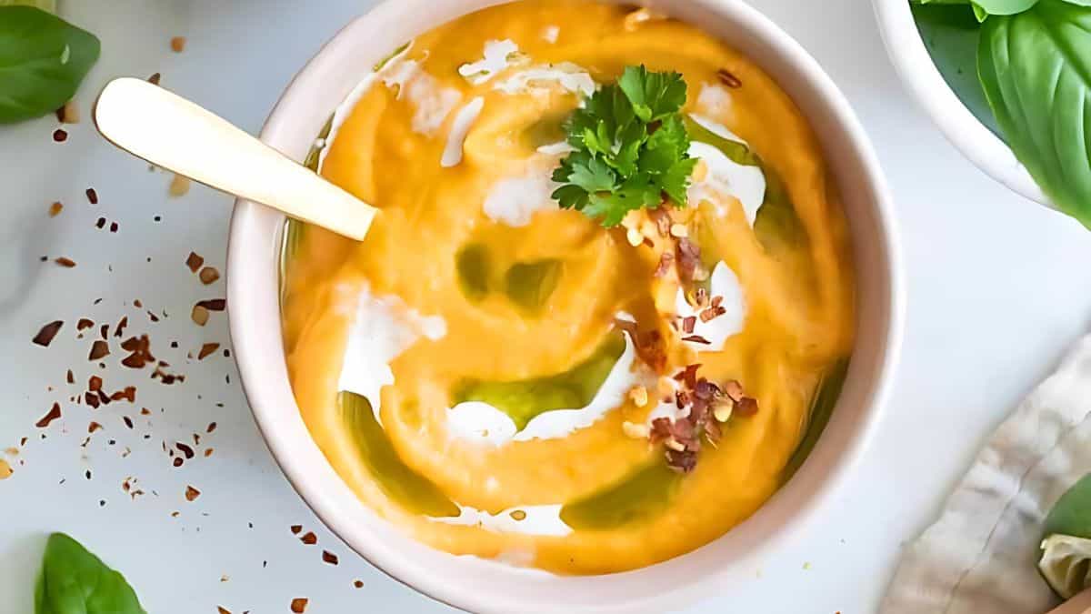 A creamy bowl of pumpkin soup swirled with coconut cream and olive oil, garnished with a sprig of fresh parsley and a pinch of red pepper flakes, with a gold spoon resting in the bowl. The soup is surrounded by fresh basil leaves on a white surface.