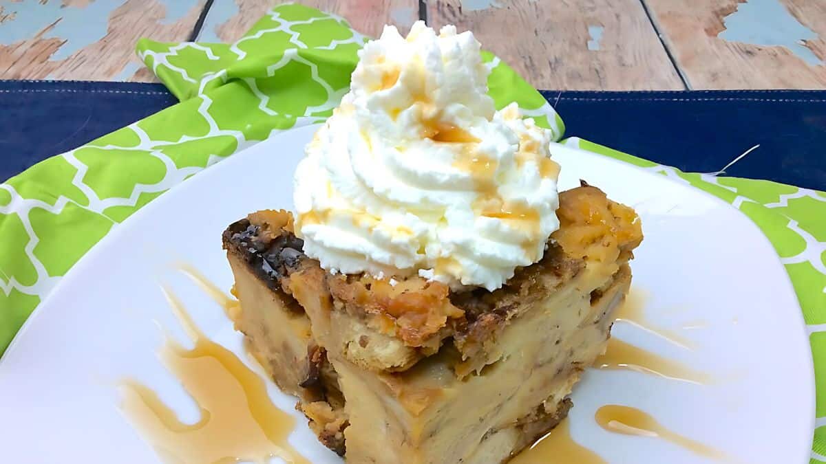 A slice of bread pudding topped with whipped cream and drizzled with caramel sauce sits on a white plate. The dessert is placed on a wooden table with a green patterned cloth in the background.