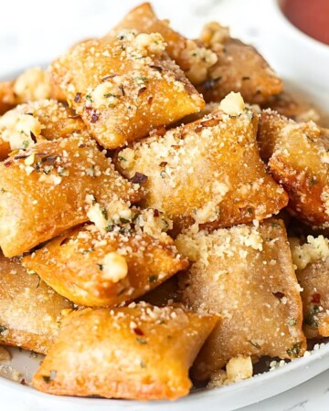 A plate of golden-brown fried ravioli topped with grated cheese is served on a white surface, making for perfect after school snacks. In the background, there is a small bowl of marinara sauce and a metal container holding crushed red pepper flakes.