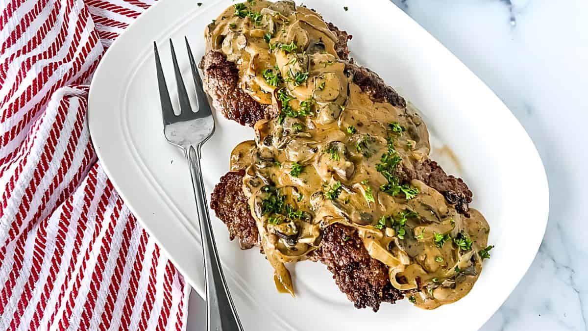 A white platter with two steaks covered in creamy mushroom sauce, garnished with chopped parsley. A metal fork lies on the side. A red and white striped cloth is partially visible.