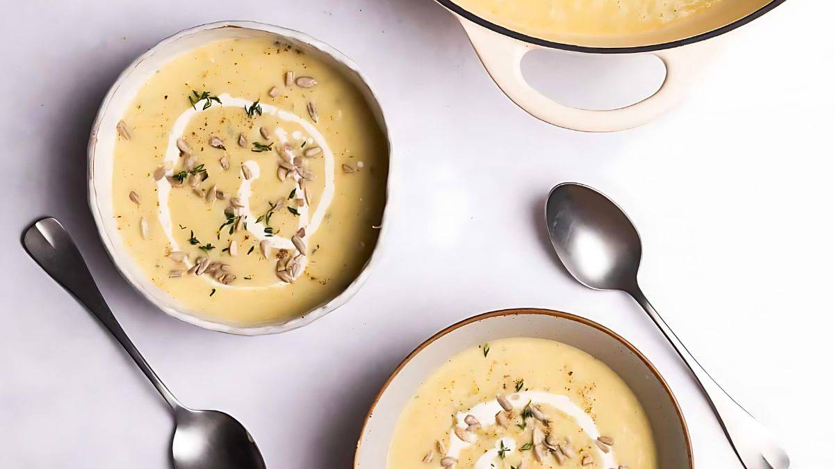 Two bowls of creamy soup garnished with a swirl of cream, herbs, and sunflower seeds. The bowls are accompanied by two spoons, and a pot of soup is partially visible at the top right corner of the image. The background is a plain white surface.