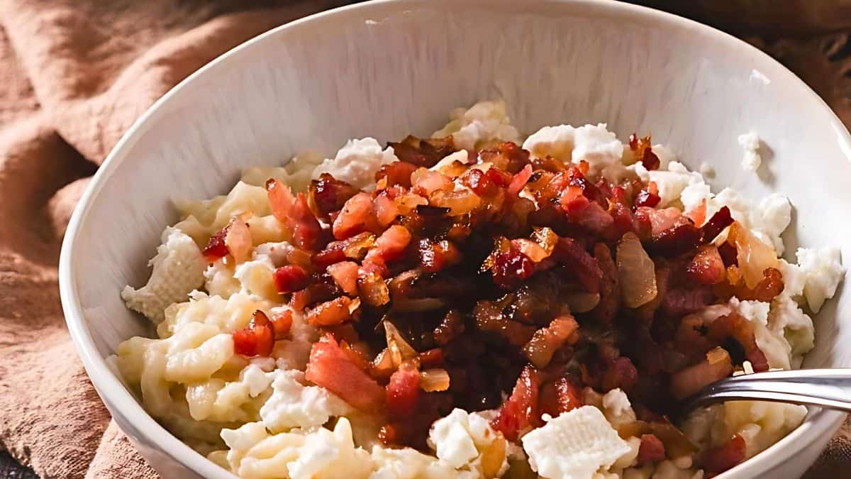 A bowl filled with a creamy dish topped with crispy, diced bacon pieces. The bacon appears golden-brown and scattered over a white, lumpy base, possibly cottage cheese or mashed potatoes. A spoon is partially visible in the bowl, and a brown cloth napkin is in the background.