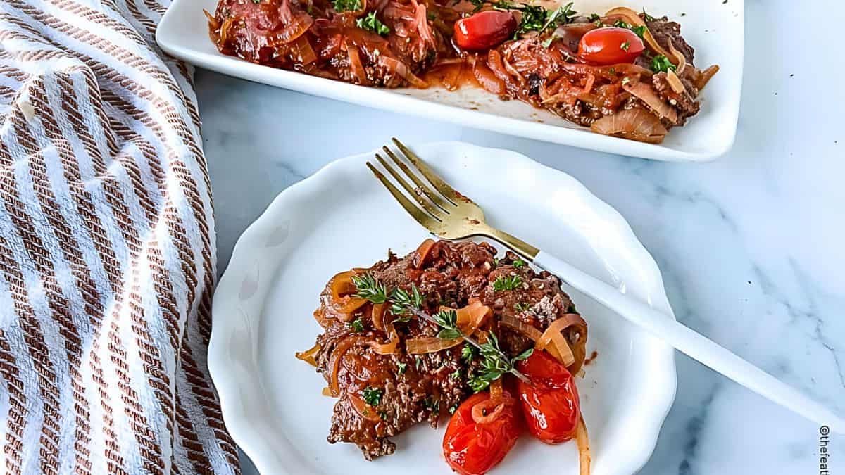 A plate of beef liver with caramelized onions, grape tomatoes, and herbs, served with a golden fork. A striped cloth is on the side, and more of the dish is visible in a serving platter in the background.