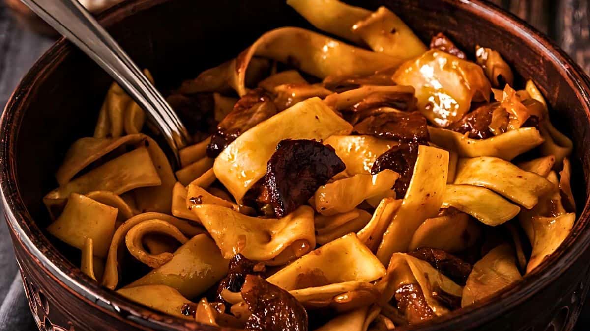 A close-up of a rustic dish featuring wide, flat noodles mixed with pieces of succulent meat in a rich, savory sauce. The dish is served in a deep, dark bowl with a metal spoon partially visible, ready to serve. The colors are warm and inviting.