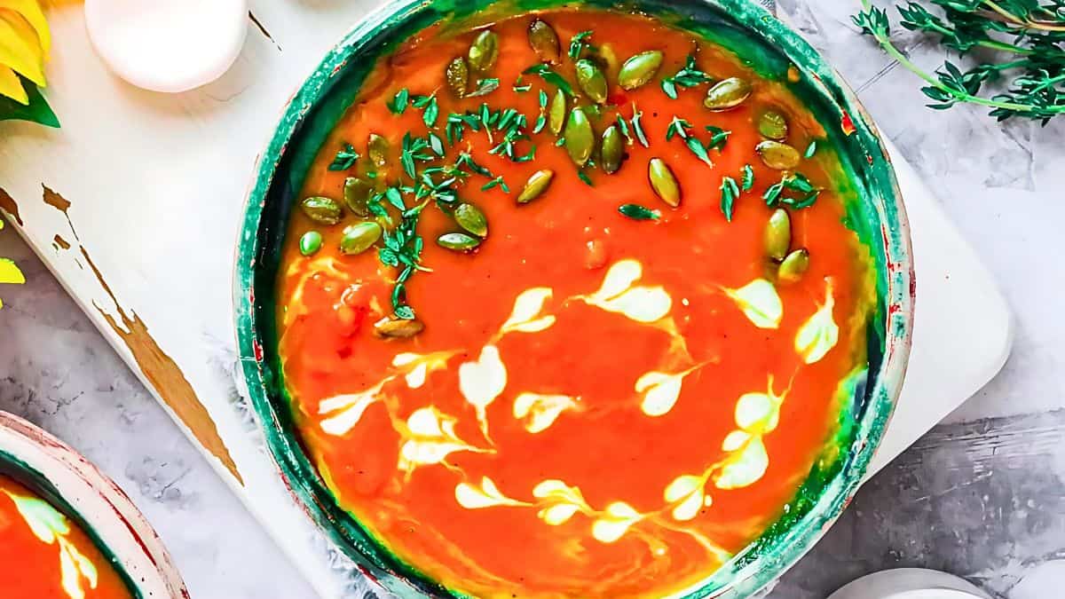A vibrant bowl of pumpkin soup garnished with a swirl of cream, fresh herbs, and pumpkin seeds. The bowl sits on a light-colored surface, accompanied by another partial bowl of soup and sprigs of fresh thyme in the background.