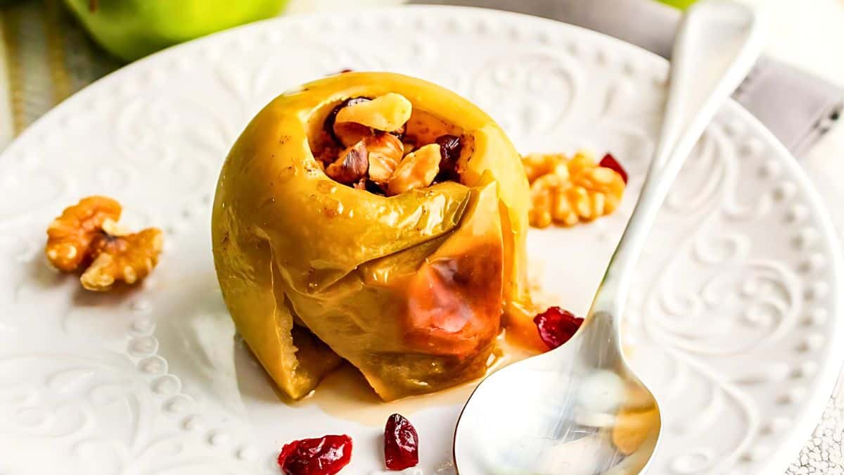 A baked apple filled with walnuts and cranberries is served on a white decorative plate. A silver spoon is placed beside it, and scattered cranberries and walnuts garnish the plate.