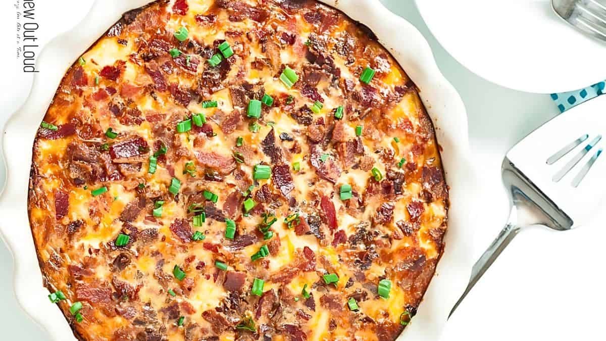 A baked dish in a round white pie dish is topped with melted cheese, crispy bacon bits, and chopped green onions. To the right, a metal serving spatula rests on a white surface, with a white plate partially visible nearby.