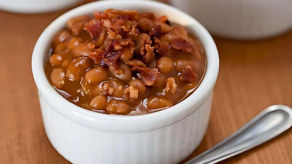 A small white ramekin filled with baked beans topped with crispy bacon bits, placed on a wooden surface. A silver spoon is positioned near the ramekin.