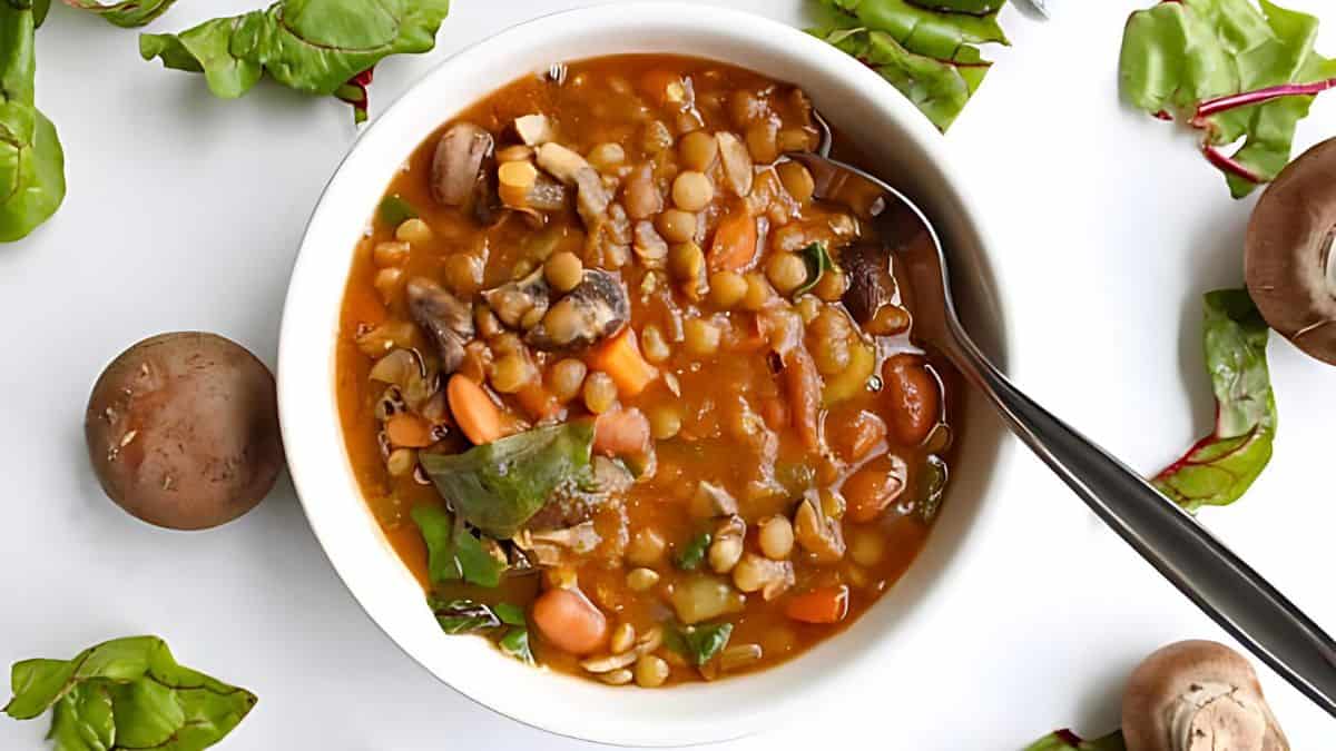 A bowl of vegetable soup containing lentils, mushrooms, carrots, and greens, placed on a white surface. Fresh mushrooms and green leaves are scattered around the bowl. A spoon is partially submerged in the soup.