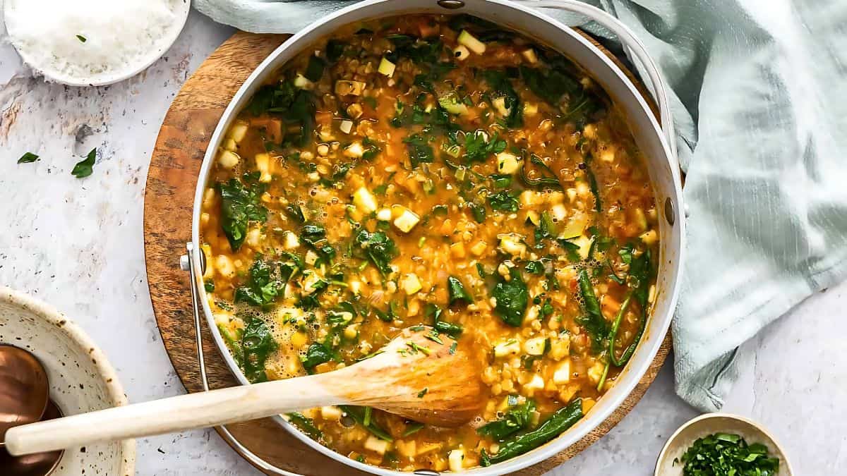A top-down view of a pot filled with a colorful vegetable stew, featuring chopped greens, diced vegetables, and a rich broth. A wooden spoon is stirring the mixture. Surrounding the pot are a wooden board, a green cloth, and various small bowls with ingredients.